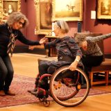 1 woman with cane stands in a semi-crouched position and reaches out to woman seated in wheel chair.