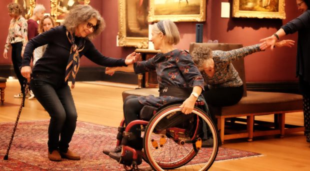 1 woman with cane stands in a semi-crouched position and reaches out to woman seated in wheel chair.