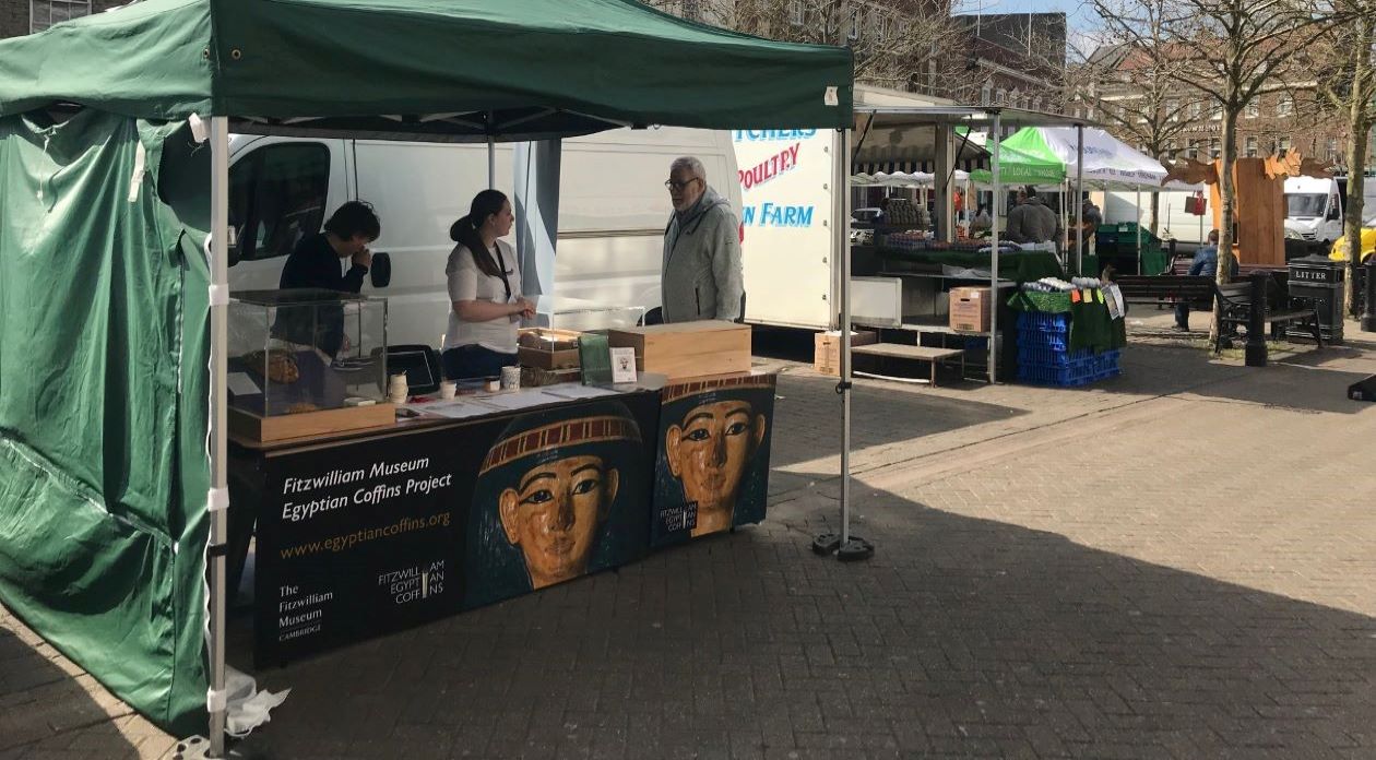 3 people stand by a market stall displaying information and Egyptian artefacts. A sign reads, 'Fitzwilliam Museum Egyptian Coffins Project'.