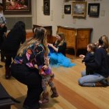 A group of adults and their young children seated on the floor of a gallery looking at paintings together