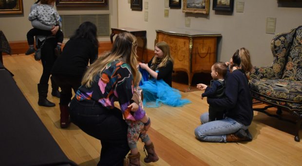 A group of adults and their young children seated on the floor of a gallery looking at paintings together