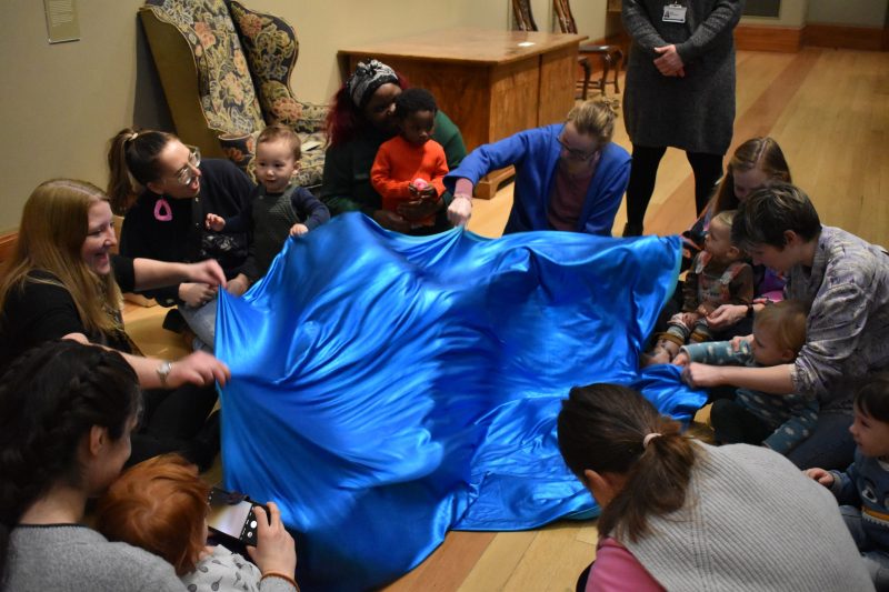 Group of adults and toddlers sit around a large piece of blue fabric on the floor which they are holding and moving.