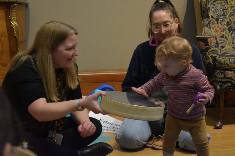 Two women are seated on the floor of a gallery. One woman holds out a tambour to a toddler who is standing touching this.