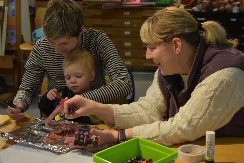 Two women and a small child sit at a table using craft materials.