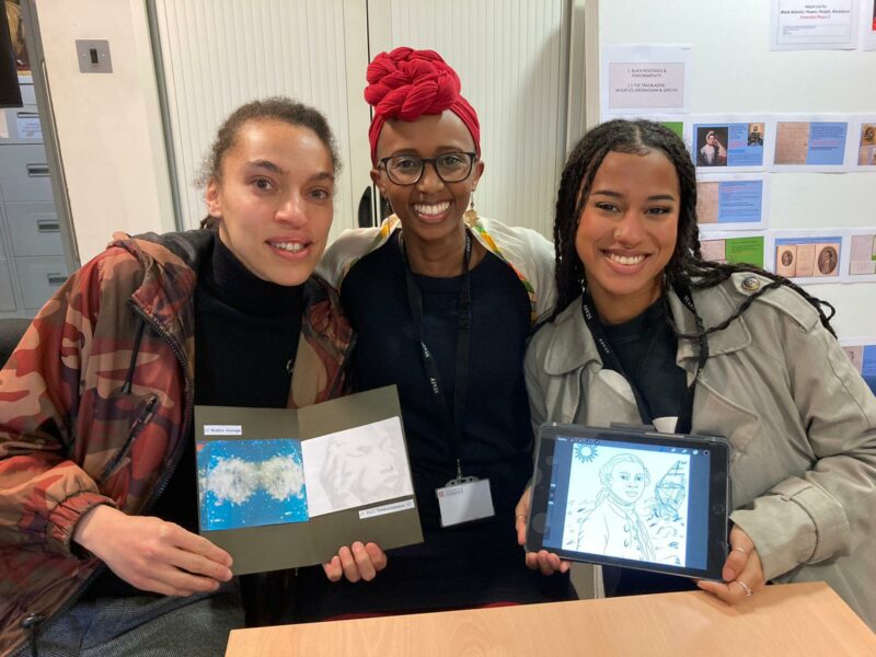 3 women of colour stand together smiling for the camera. 2 of the women are holding examples of their art work