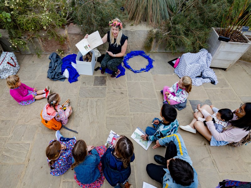 A woman wearing a flower garland and holding an open storybook, kneels in front of a circle of seated children.