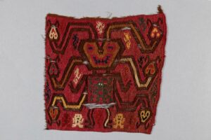 Embroidered woollen textile fragment with a human-like figure with six radiating serpents. The rich red background is from an insect-based dye known as cochineal.