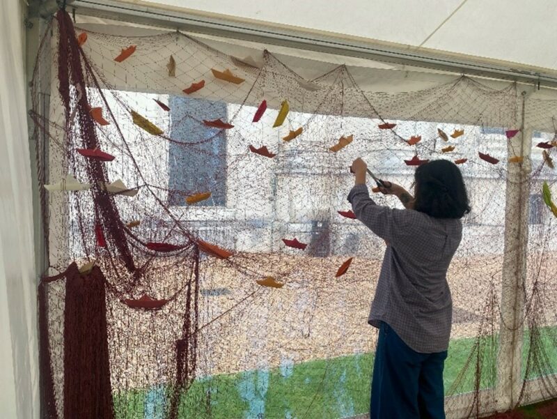 A woman with back to the camera attaches coloured paper boats to a hanging fishing net