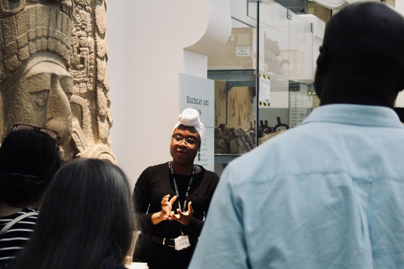 A woman of colour stands beside a carving in front of a group of people.