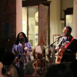 Man holding guitar stands in front of microphone in a museum gallery