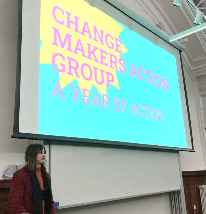 Woman sits underneath large projection screen which reads: Change Makers Action Group Year of Action