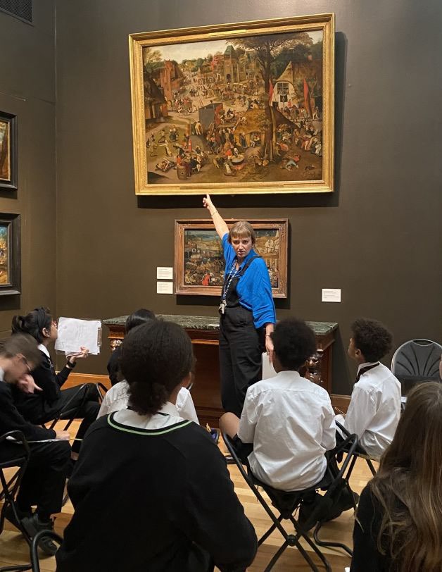 A woman stands and points at a large, detailed painting in a gilded frame in front of several children sitting on stools