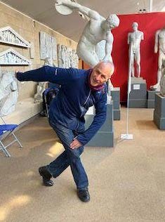 A white man stands in front of a plaster cast of a discus thrower, copying the pose