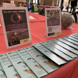 A table with a red tablecloth set up for the Cambridge City Council holiday lunch activity session. There is a line of museum leaflets and two flyers on stands about the Whipple Museum.