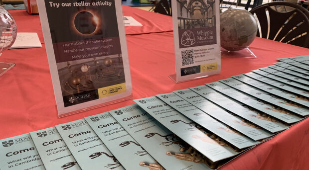 A table with a red tablecloth set up for the Cambridge City Council holiday lunch activity session. There is a line of museum leaflets and two flyers on stands about the Whipple Museum.