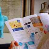 Someone holds open a Museum Walk leaflet in front of a turquoise hawk sculpture.