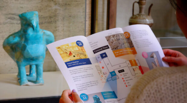 Someone holds open a Museum Walk leaflet in front of a turquoise hawk sculpture.