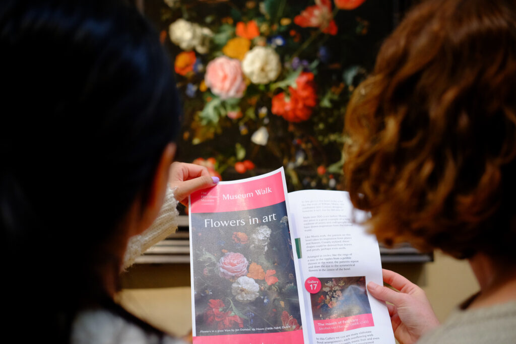 Two people seen from behind holding a Flowers in Art Museum Walk leaflet, in front of a painting of some flowers at the Fitzwilliam Museum.
