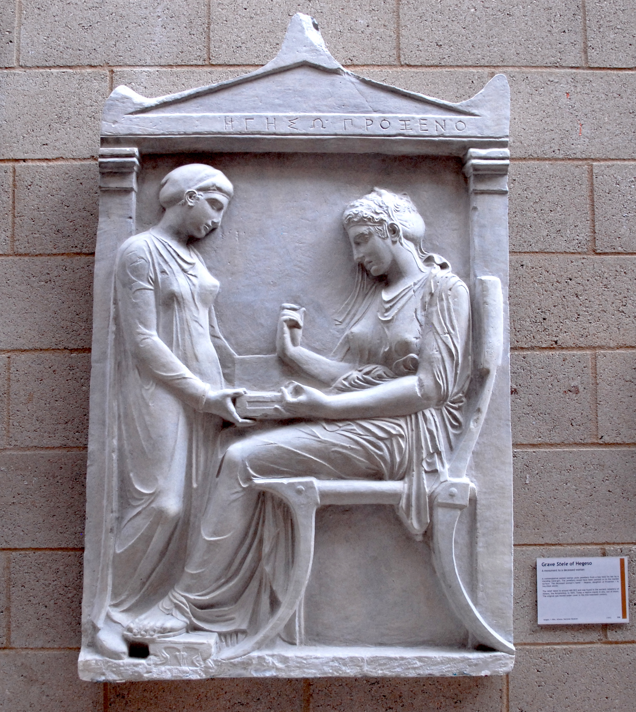 Plaster cast of Hegeso's grave monument, which is roughly rectangular, with a triangular pediment where Hegeso's name is written in Greek. It shows two women: on the right, a woman sits on a chair facing left, wearing a long, draped dress and elaborate hairstyle; opposite her stands a smaller woman in plain clothes, holding out a box. The seated woman's left hand holds the box too. Her feet rest on a small footstool.