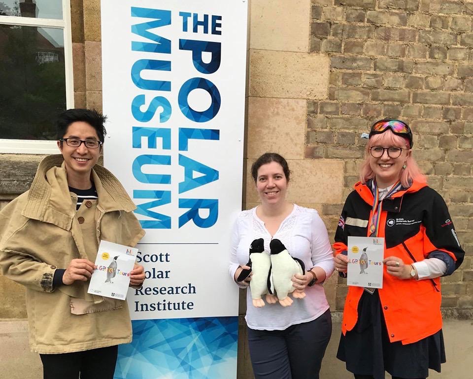 Dan Vo, Charlotte Connelly and Ellie Armstrong outside the Polar Museum. Behind them is a large banner saying "Polar Museum". Dan and Ellie are holding Bridging Binaries flyers, while Charlotte holds two cuddly penguins.