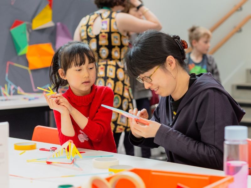 Young girl in red jumper and adult woman in blue top and glasses sitting at table doing a craft activity with coloured straws