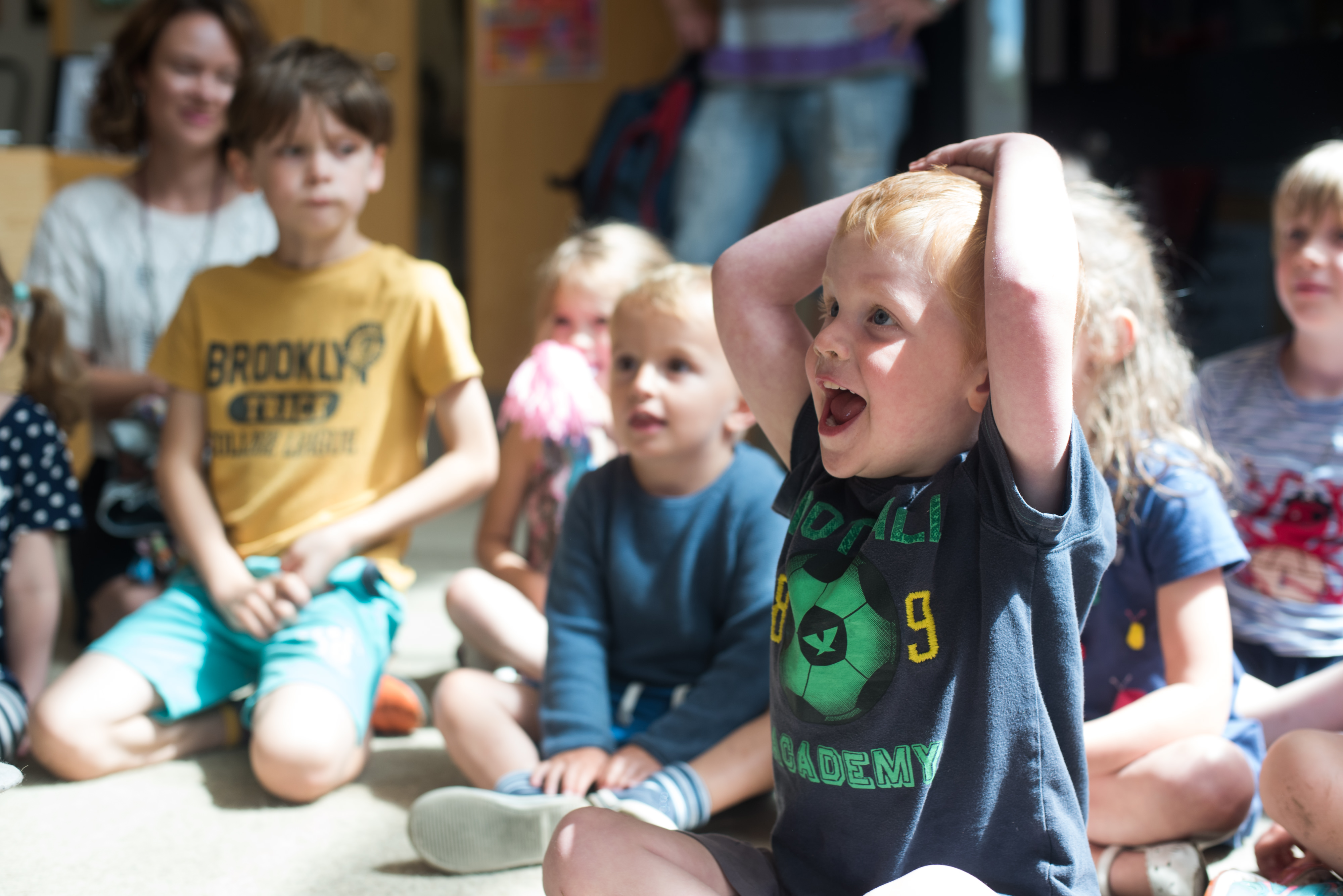 Group of children sitting on the floor, with one child at the front smiling with his hands on his head.