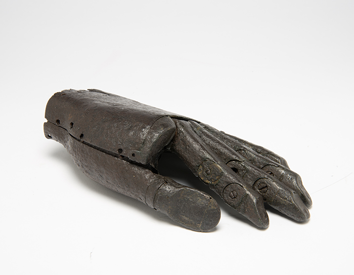 iron prosthetic hand side view. It is a left hand, and lies palm down with the thumb underneath. The hand ends at the wrist and is made of dark iron and the four fingers are fully articulated, while the thumb is not jointed