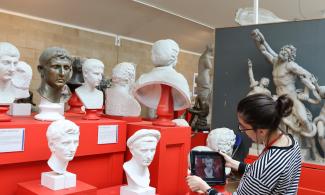 Person standing in front of busts of roman emperors inside the museum. She is holding a iPad. Laocoon in the background.