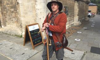 Man dressed as a 17th century soldier wearing black hat, red jacket and leaning on a cane on a street corner