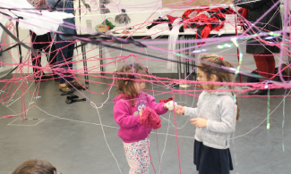 Two children surrounded by strings of yarn stretched across the room/
