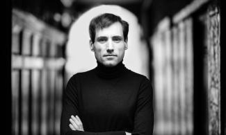A black and white photo of a man looking at the camera with his arms folded, wearing a black turtleneck