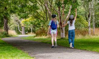 Two young girls walk along a path through grass and trees. One points up at one of the trees.