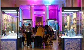 A group visitors gather around a table, with brightly coloured yellow chairs, in the centre of a museum gallery