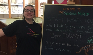 A museum tour guide smiling enthusiastically next to her tour advert, on a museum chalk board