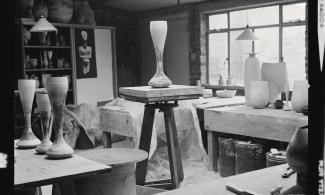 A black and white photograph of a ceramic artist's studio, with a central long, thin, circular ceramic piece on top of a trestle table.