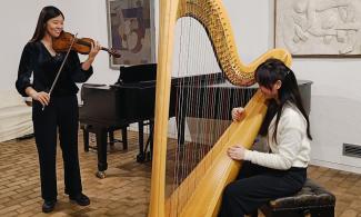 Two students playing a concert in Kettle's Yard house, one is standing, playing the violin, and the other sits playing the harp.