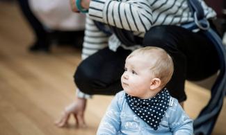 alt='a baby is sat on the floor in a blue shirt, his mother crouches behind"