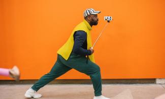 A man wearing green trousers lunges in front of a bright orange wall, holding a selfie stick.