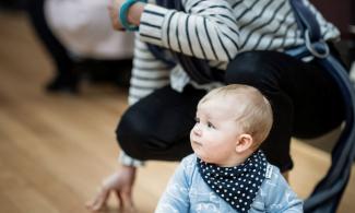 alt='a baby is sat on the floor in a blue shirt, their mother crouches behind"