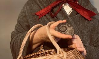 An actress playing the palaeontologist Mary Anning. She is wearing a shawl and the red ribbon of her bonnet is visible, her face is not in the picture. She is holding a basket and an ammonite fossil.. 