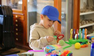 Young boy in blue hat plays with an assortment of sensory toys laid out on a table before him. Museum cases are in the background. 