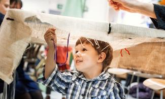 alt="a young child with blond hair and a blue checkered shirt plays with string and cloth in the museum"