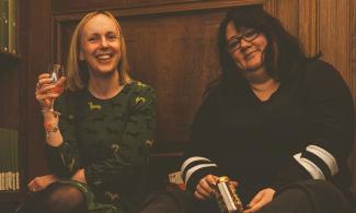 Two women smiling and sitting in a wood panelled room in the University Library each holding a drink, one a glass of wine, the other a can.