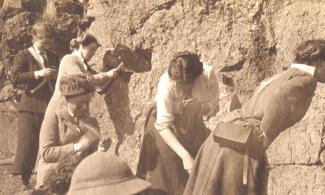 A sepia photograph of 5 female, early geologists studying a rock - face wall, some tapping the wall and rocks with geological hammers. 