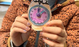 A child’s glittery purple-painted hands hold up a handmade pocket watch with a pink face and purple and blue hands. The child is wearing a brown coat with dark spots.