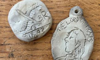 Two clay medal models sit on a wooden surface. The left medal has dates and patterns carved into it; the right features a carved side profile of a man and a hole at the top.