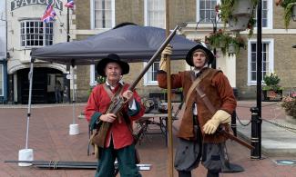 Two men stand in a town square dressed as Civil War soldiers. The man on the left holds a large gun, while the man on the right carries a long pole. A gazebo is visible behind them.