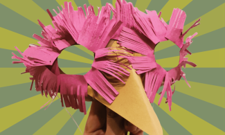 A hand holds up a handmade bird mask featuring a long yellow beak and bright pink paper feathers cut into slits around the eyes. The background is light and dark green stripes.