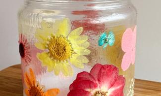 A transparent jar sits on a wooden surface, decorated with pressed flowers in shades of red, pink, orange, yellow, and blue.