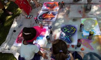 A sheet on the floor is covered in artwork and spray cans. Children and adults gather around, actively contributing to the their piece.
